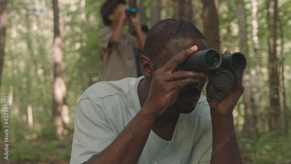 Medium close-up of concentrated Black man watching forest birds and animals through binoculars having summer camping trip with his little sons helping him