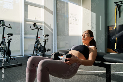 young latina woman concentrating on performing hip thrust with a dumbbell alone in gymnasium