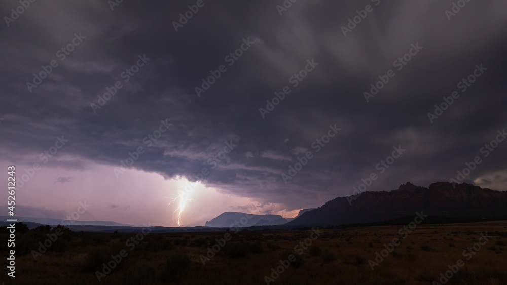 A bolt of lightning drops down from the edge of rolling purple clouds in a landscape with an open grassy field dotted with sagebrush and the mountains of Southern Utah in the distance. 