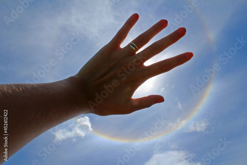 Woman's hand with ring and painted nails covering the sun in a solar halo effect, on a celestial sky.