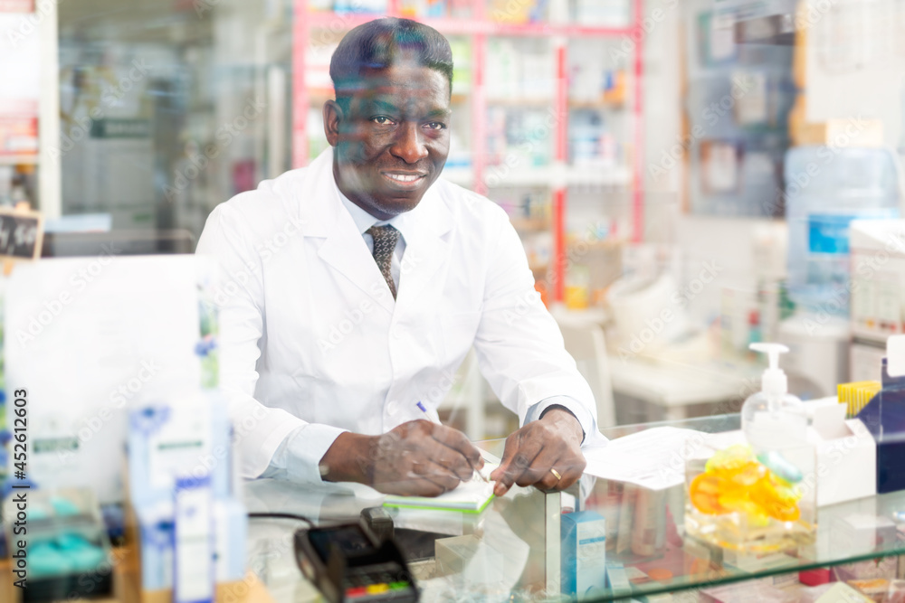 Smiling african american male pharmacist working in a pharmacy makes important notes while sitting behind the counter