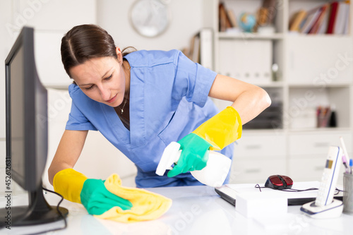 Cleaning lady of cleaning company wipes dust from table in the office