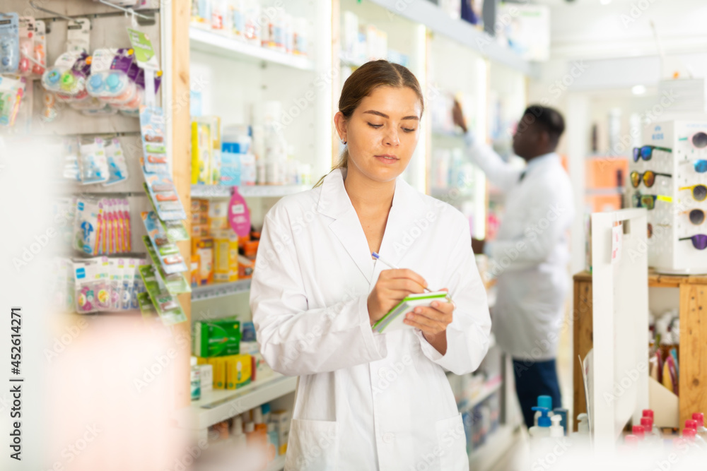 Portrait of a focused young female pharmacist standing in the sales hall of a pharmacy, making important work notes in a ..notebook