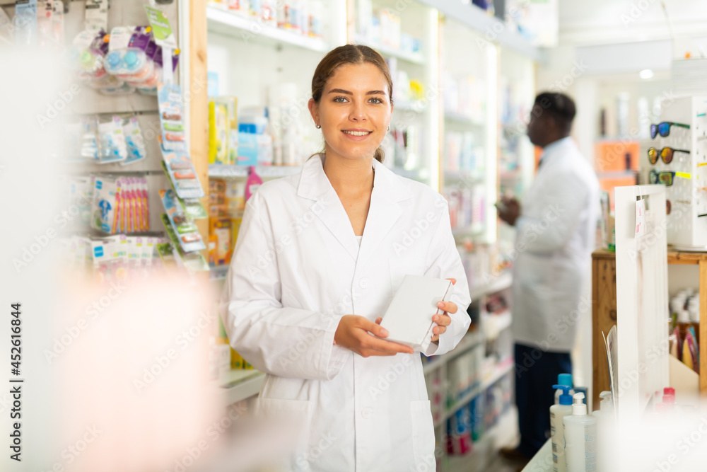 Female pharmacist standing with drug package in salesroom of drugstore. Her colleague standing behind.