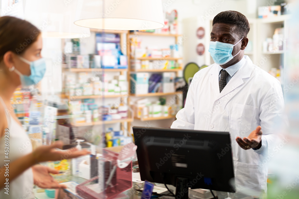 African-american male pharmacist in mask standing behind counter and talking with client