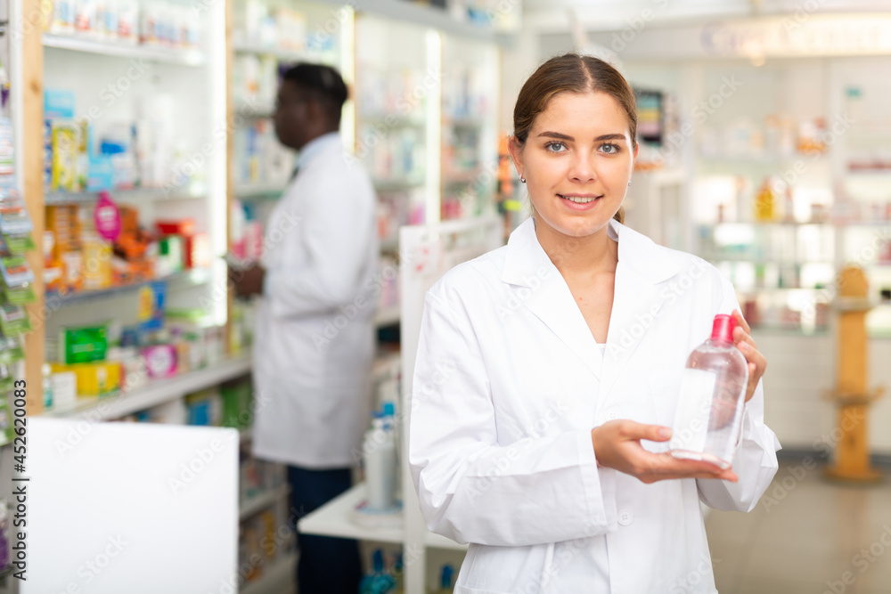 Portrait of a smiling young female pharmacist in the sales hall of a pharmacy, demonstrating recently received goods for ..sale in hands
