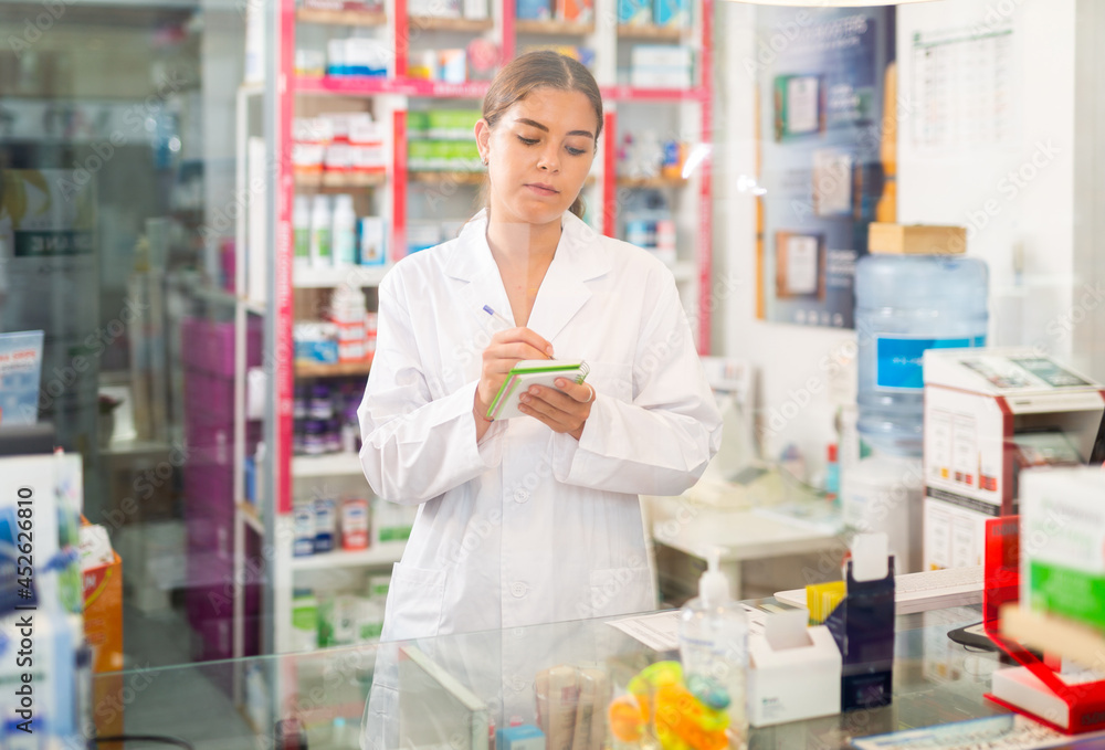 Female pharmacist in gown standing behind counter in drugstore. She's writing recipe.