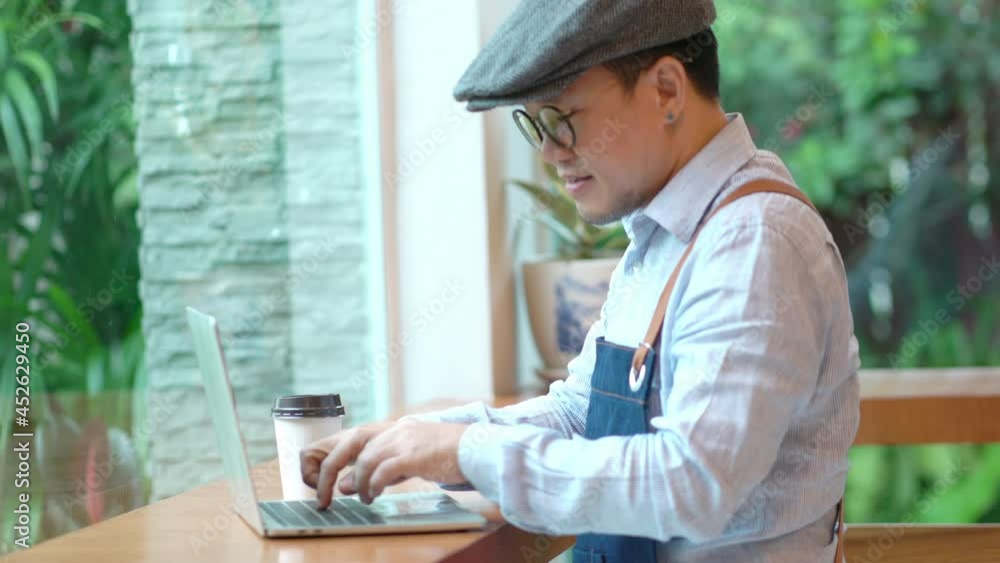 Asian man coffee shop owner sitting at table and calculating shop finance bill on laptop computer after closed. Stressed male small business restaurant entrepreneur counting payment and income bills. Asian man coffee shop owner sitting at table and calculating shop finance bill on laptop computer after closed. Stressed male small business restaurant entrepreneur counting payment and income bills.