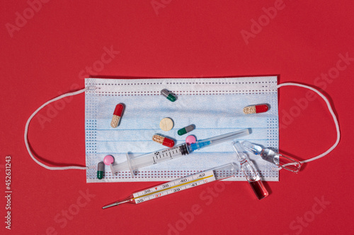 Syringe, thermometer and medicines with pills to lower the temperature lie on a face mask on a red background, top view