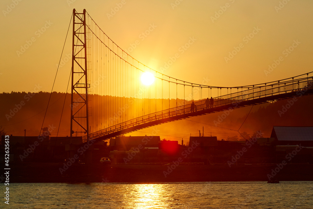 People walk on a suspension bridge over the river at sunset