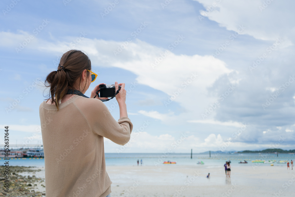 back view of woman wearing sunglasses standing on the beach take a photo by digital camera with sunny day
