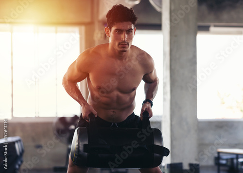 Bilde på lerret Muscular sporty young man working out with heavy sandbag while exercises in the gym
