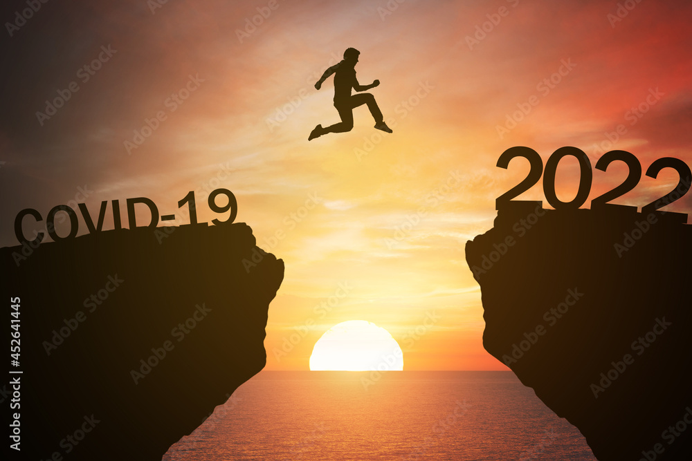 New year concept; pass from covid-19 to 2022 , silhouette man jump over barrier cliff and success with beautiful sunrise with sea background. Starting 2022 with goals and success. Use for banner. New year concept; pass from covid-19 to 2022 , silhouette man jump over barrier cliff and success with beautiful sunrise with sea background. Starting 2022 with goals and success. Use for banner.