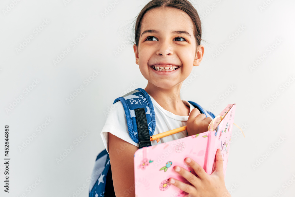 Closeup portrait of a happy smiling schoolgirl with blue backpack writes something on her notebook, looking at one side during posing over light grey studio background. Closeup portrait of a happy smiling schoolgirl with blue backpack writes something on her notebook, looking at one side during posing over light grey studio background.