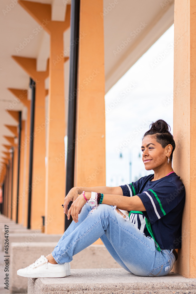 Mid adult afro mexican woman wearing blue ripped jeans, sitting and leaning against a concrete column