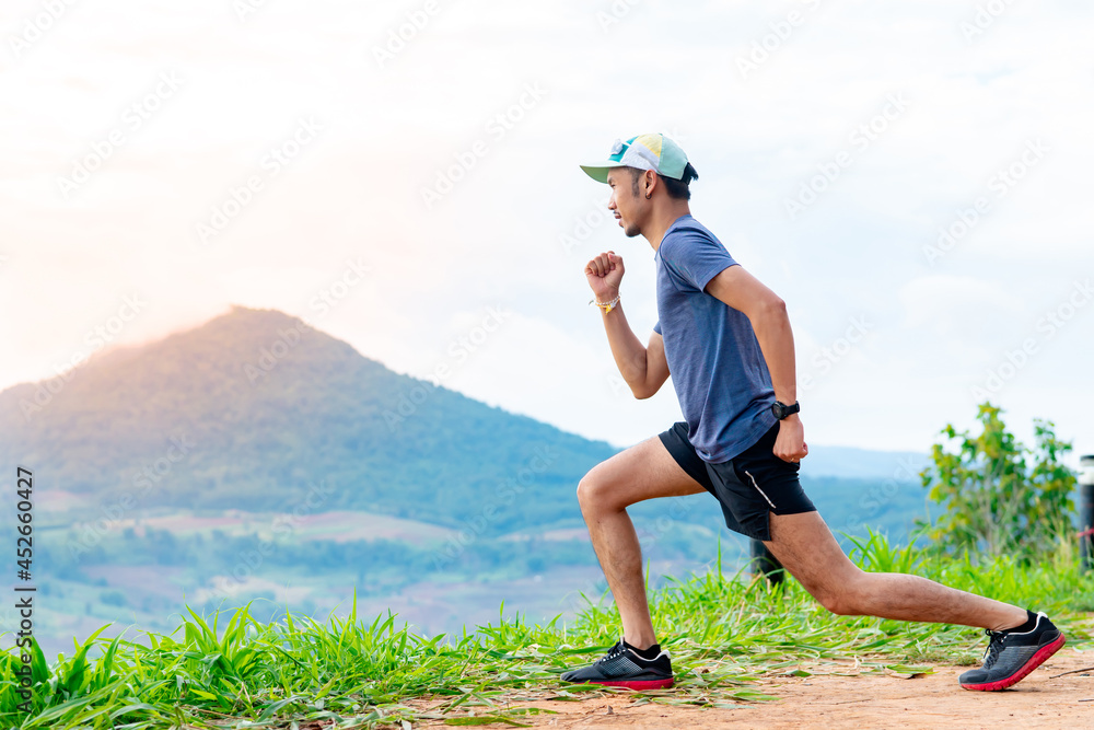 Portrait of an Asian male runner in a blue shirt and hat standing warm up in preparation for a jogging run. It is located on a high cliff with beautiful mountains in the background. Portrait of an Asian male runner in a blue shirt and hat standing warm up in preparation for a jogging run. It is located on a high cliff with beautiful mountains in the background.