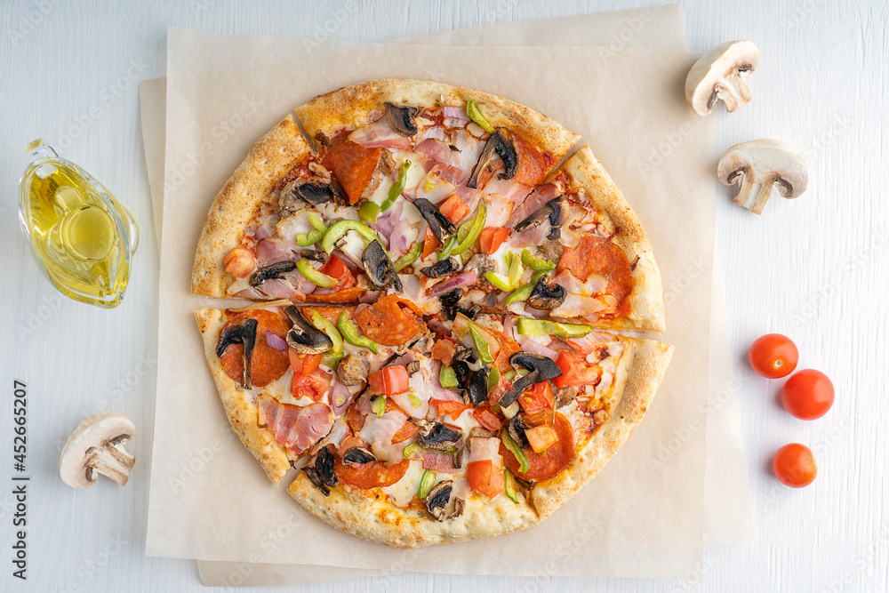 Top view of one whole sliced tasty italian pizza made of mushrooms, tomatoes, ham, pepper and cheese served on craft brown paper with olive oil on white wooden background. Horizontal image