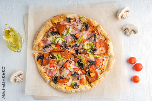 Top view of one whole sliced tasty italian pizza made of mushrooms, tomatoes, ham, pepper and cheese served on craft brown paper with olive oil on white wooden background. Horizontal image