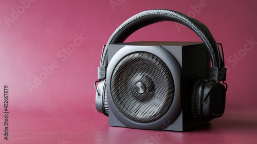 Black single-band speaker and headphones with ear pads on a dark red background. Wireless audio concept. Free space for an inscription