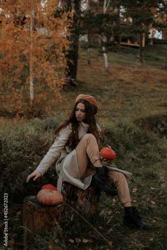 A girl in a gray coat with three orange pumpkins sits on the stump in the fall.