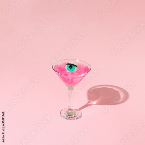 Eyeball figurine with eyelash in martini cocktail glass on pastel pink background. Halloween minimal creative concept. Blue eye retro fashion aesthetic or cosmetic idea.