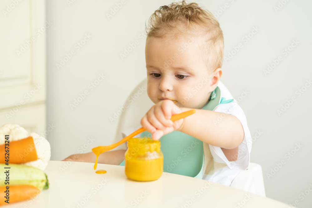 funny baby boy in bib eating vegetable puree with spoon sitting in child chair. Child learn to eat by himself funny baby boy in bib eating vegetable puree with spoon sitting in child chair. Child learn to eat by himself