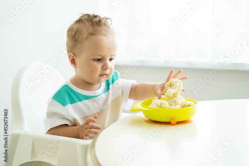 smiling baby boy sitting in a Childs chair eating cauliflower on white kitchen.