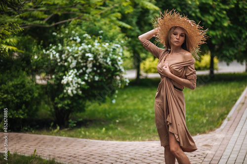 A woman in a long brown dress and a straw hat walks in a summer park