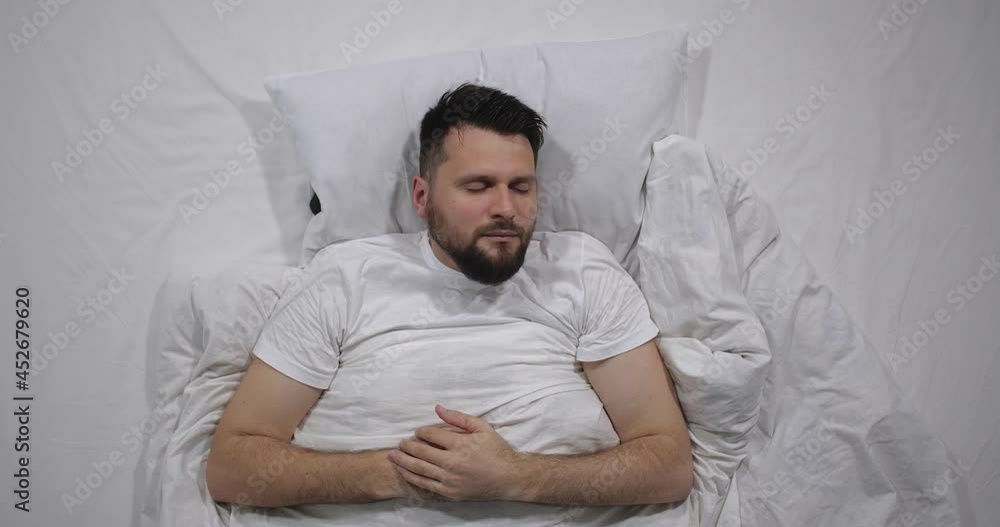Top view of caucasian bearded man was woken up by night call bed with white linens in light room. High angle shooting. Attractive guy is happy that somebody calls him 4k Top view of caucasian bearded man was woken up by night call bed with white linens in light room. High angle shooting. Attractive guy is happy that somebody calls him 4k