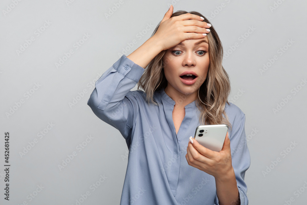 Naklejka premium Shocked young woman read stressful or surprising message in smartphone holding hand on forehead worried with mouth open in shock. Isolated studio shot of amazed girl get email or text in mobile phone