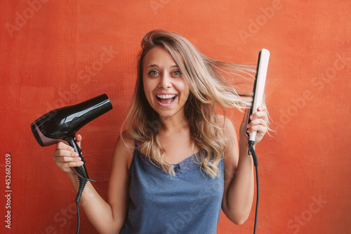 A young caucasian charming blonde woman with wavy hair in home clothes holds a hair dryer and a straightener in her hands standing against an orange wall. Hair styling, damage