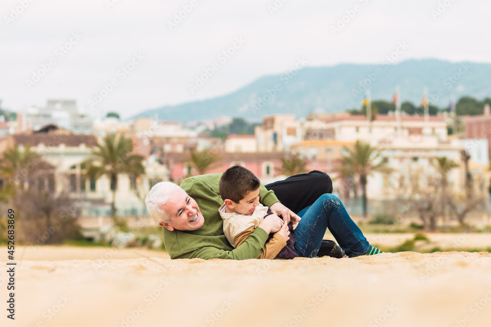 Funny boy and his grandfather playing stretched out in the sand on the beach