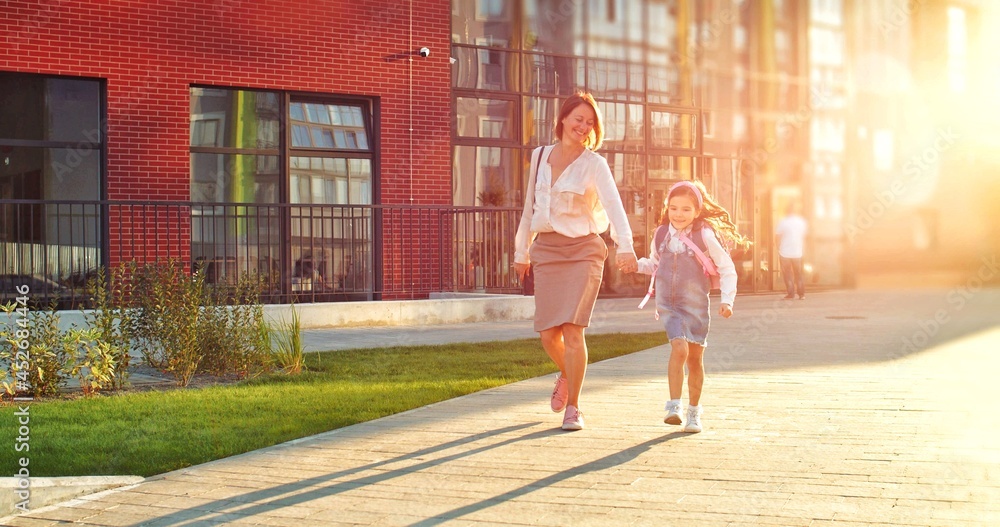 Caucasian happy mother and cute daughter walking outdoors on sunny day from school after lessons. Pretty joyful woman with little schoolgirl jumping and smiling on street. Education concept