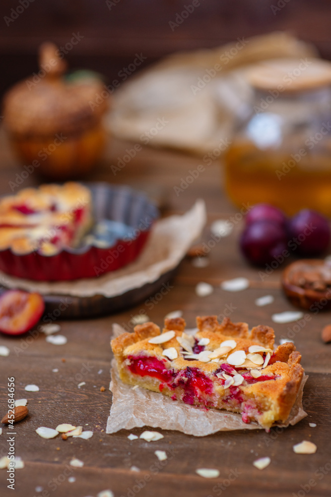 Sandy plum tarts with frangipan and almond petals on a wooden background decorated with plums, one cake is cut and the other is on the background. Sandy plum tarts with frangipan and almond petals on a wooden background decorated with plums, one cake is cut and the other is on the background.