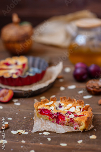 Sandy plum tarts with frangipan and almond petals on a wooden background decorated with plums, one cake is cut and the other is on the background.