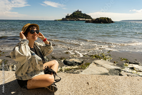 Portrait of a young female sitting on the promenade and holding her hat while witnessing St Michael´s Mount during high tide.
