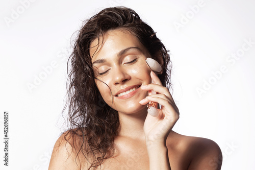 Smiling woman touching cheek by jade roller, massaging face, enjoying perfect smooth skin with closed eyes. Beautiful young girl with naked shoulders doing facial massage by guasha massager.