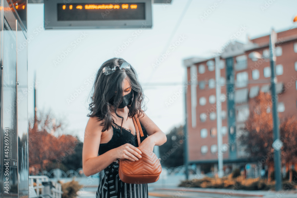 Mujer mayor morena con mascarilla revisando su bolso en la estación de tren junto a la ciudad - obrazy, fototapety, plakaty 