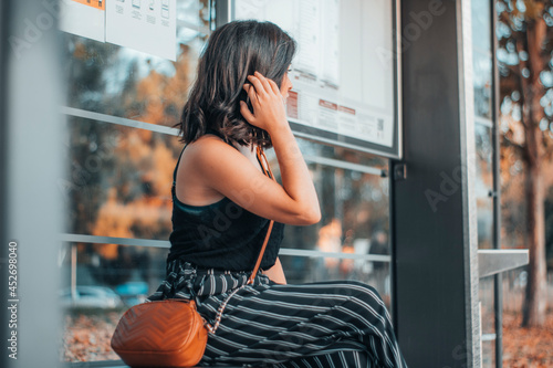 Chica adulta de pelo castaño esperando en la parada del autobús sentada para realizar el transporte publico