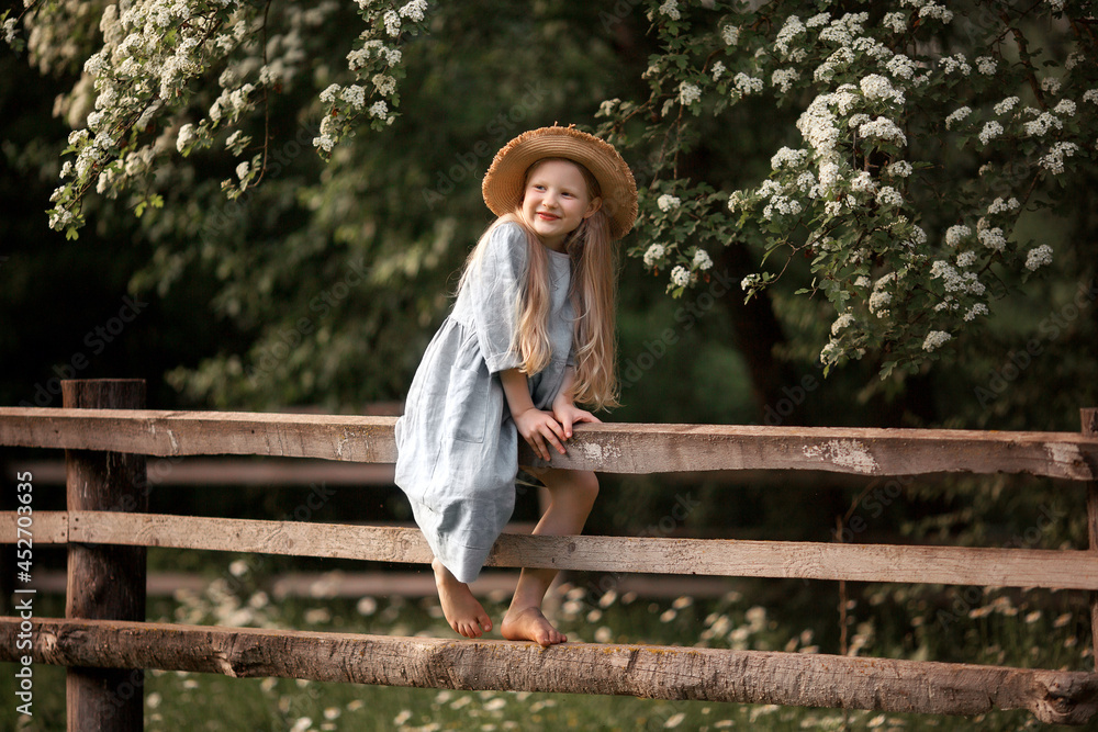 Obraz premium a little girl in a blue linen dress and a straw hat climbs fences in the village in summer