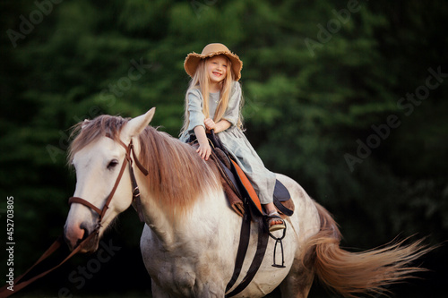 a little girl in a blue linen dress and a straw hat is stroking a horse riding © AnastasiaKharichkina