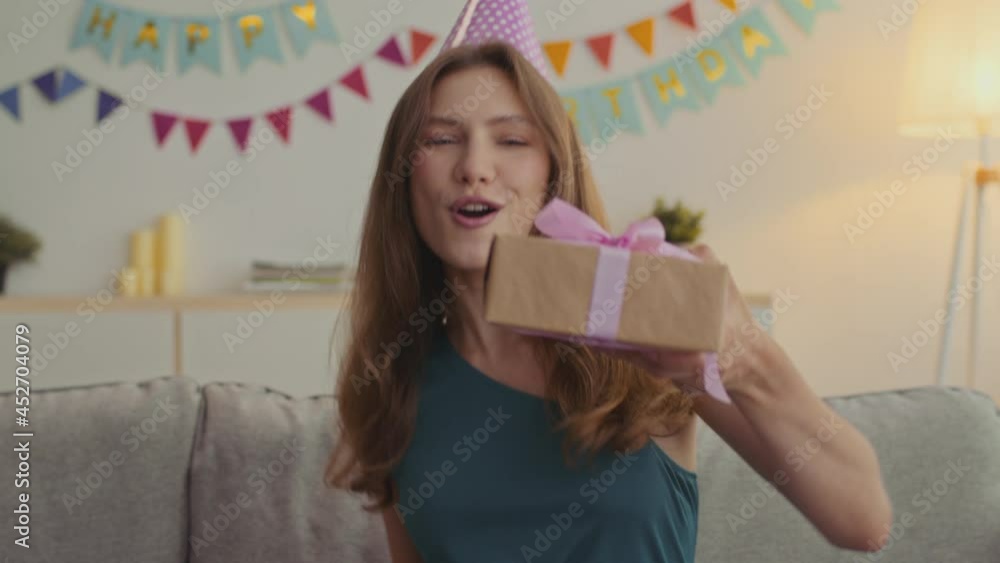 Happy birthday greeting. Excited young lady wearing holiday cap blowing up whistle and offering gift box to camera Happy birthday greeting. Excited young lady wearing holiday cap blowing up whistle and offering gift box to camera