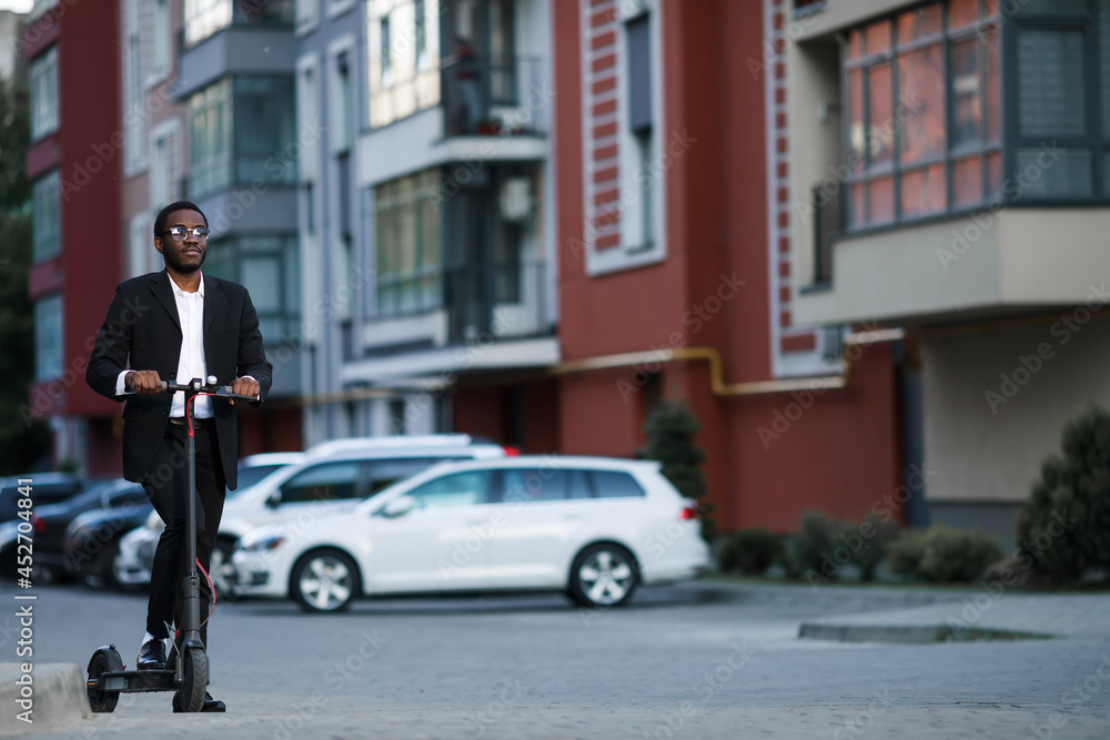 A man in a black business suit is riding an electric scooter