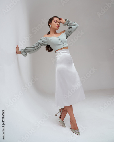 Elegant woman wearing pastel green top, silver silk skirt and high heels standing at white gray background and leaning on wall. Pretty fashion female model with pony tail brunette hair