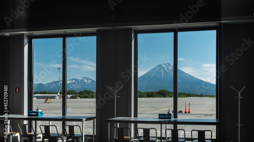 Conical volcanoes against the blue sky are visible through the airport windows. There is snow on the slopes. In the foreground are tables and chairs of the cafe. Petropavlovsk Kamchatsky
