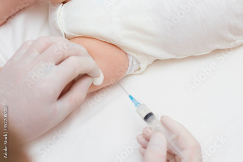 The doctor s hands in rubber protective gloves hold a syringe. An infant receiving the vaccine in the leg. Visit to the pediatrician in the hospital. A two-month-old baby. To close.