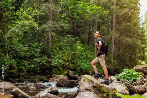 Traveler hiker with backpack enjoys landscape by mountain river in Carpathian forest. Trip to summer Ukraine