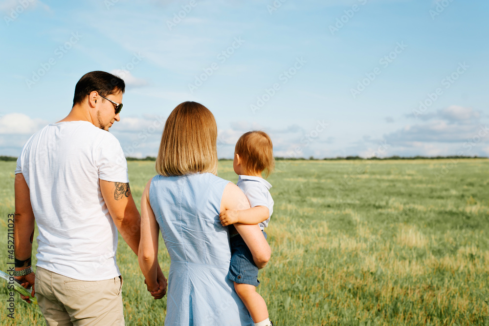 Young family with small child walk in field. Back view young father holding wife by hand, mom carries little son in her arms, couple in love spending time with each other. Family outdoor recreation
