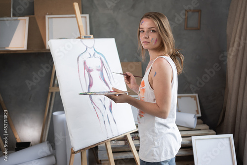 Caucasian girl artist draws a nude in a creative studio with a pencil on canvas. A talented female painter works in the workshop. the girl looks into the camera.Mature Content