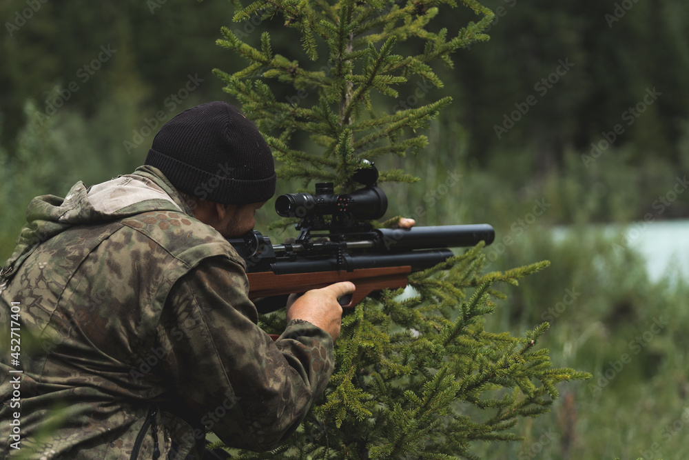 The hunter aims with a rifle. A man in camouflage is preparing to shoot. Hunting in the woods with a sniper rifle